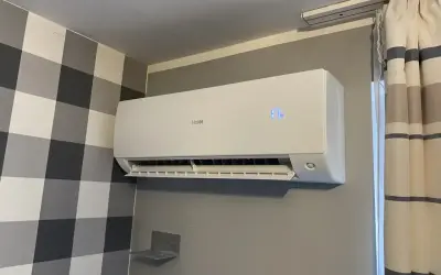 Biała klimatyzacja Haier zamontowana na szarej ścianie, obok wzorzystej tapety i zasłon.