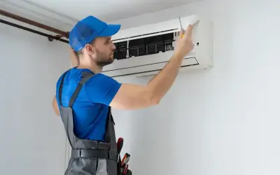 Technik w niebieskiej czapce i szarym ogrodniczkach naprawia klimatyzator śrubokrętem.