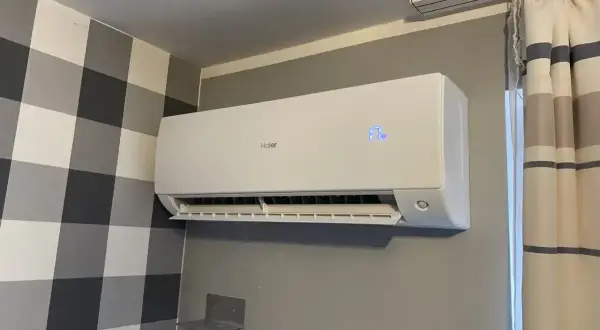 Biała klimatyzacja Haier zamontowana na szarej ścianie, obok zasłony.